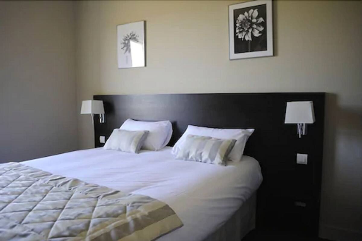 Una cama bien tendida con sábanas blancas y un edredón acolchado, dos lámparas fijadas a la pared y dos cuadros con motivos florales en la pared.