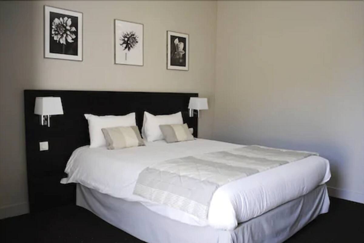 Una cama bien hecha con sábanas blancas, dos lámparas de noche y tres cuadros en la pared.