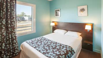 A hotel room with a bed, two wall-mounted lamps, a window with curtains, and two framed pictures on the wall.