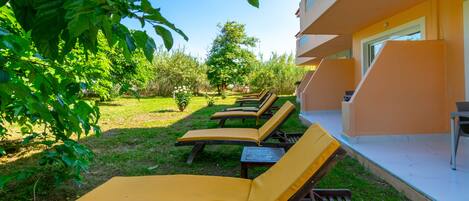 A row of wooden lounge chairs with yellow cushions in a garden area, adjacent to a building with a yellow facade.