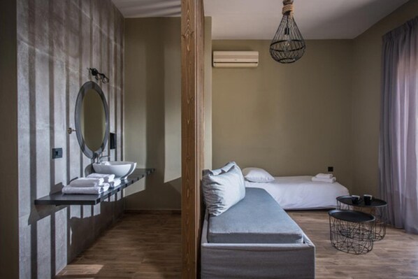 A modern hotel room with a wooden floor, a bed with white linens, a grey sofa, a round mirror above a white sink, and a hanging light fixture.