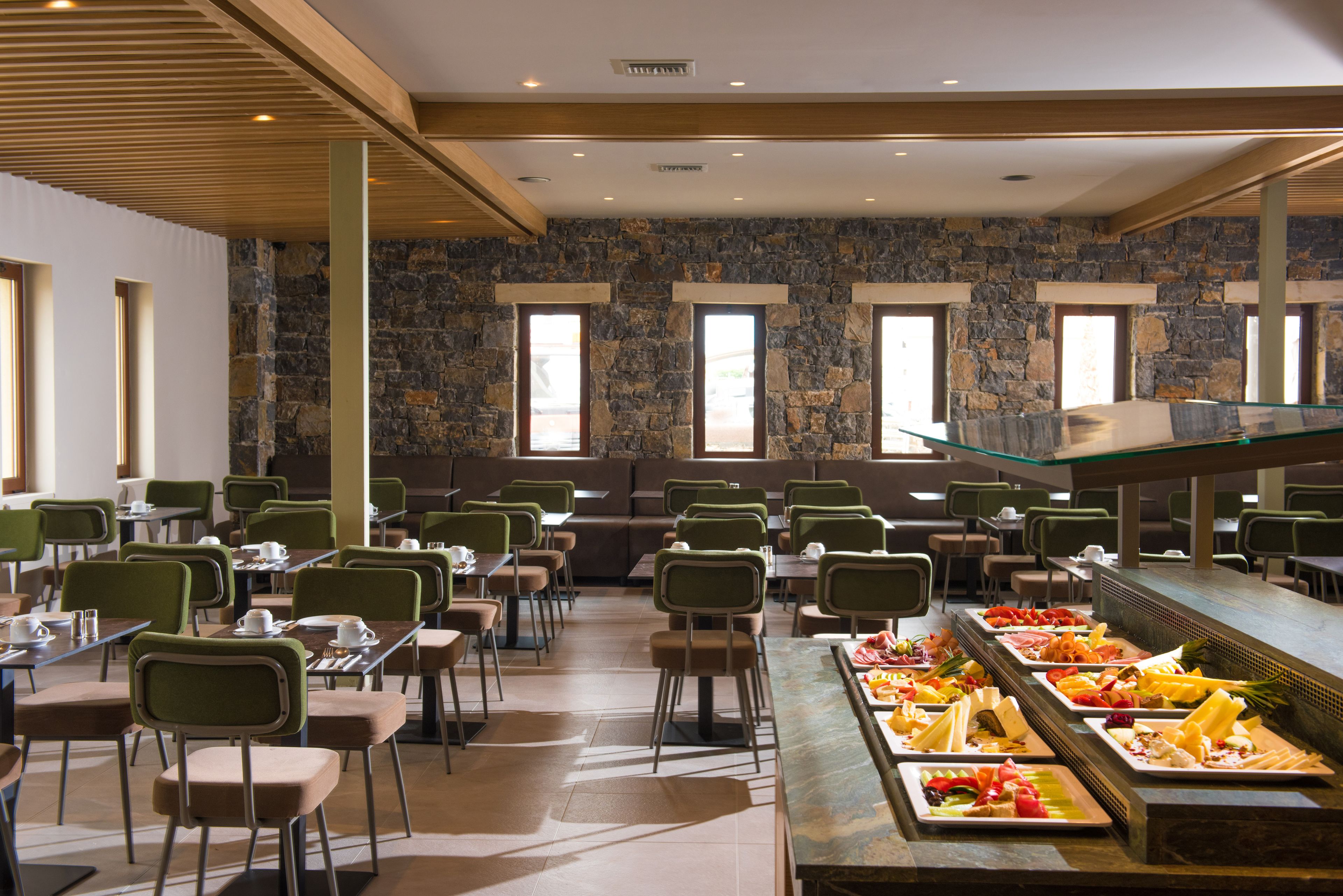 Un amplio comedor con varias mesas y sillas, un bufé con diversas opciones de comida, y una pared de piedra con ventanas.