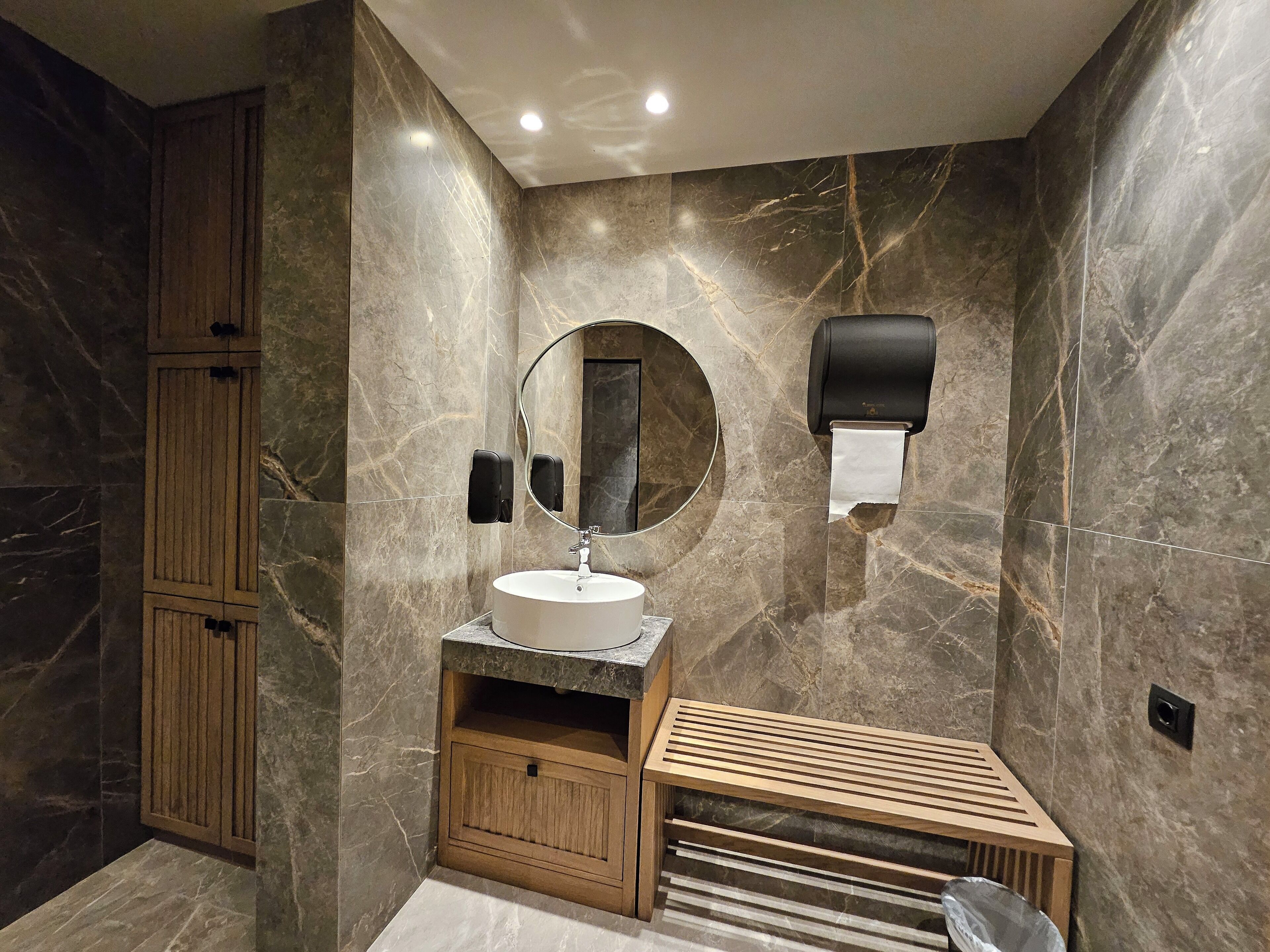 Ein modernes Badezimmer mit Marmorwand, Waschtisch aus Holz, rundem Spiegel und Toilettenpapierhalter.