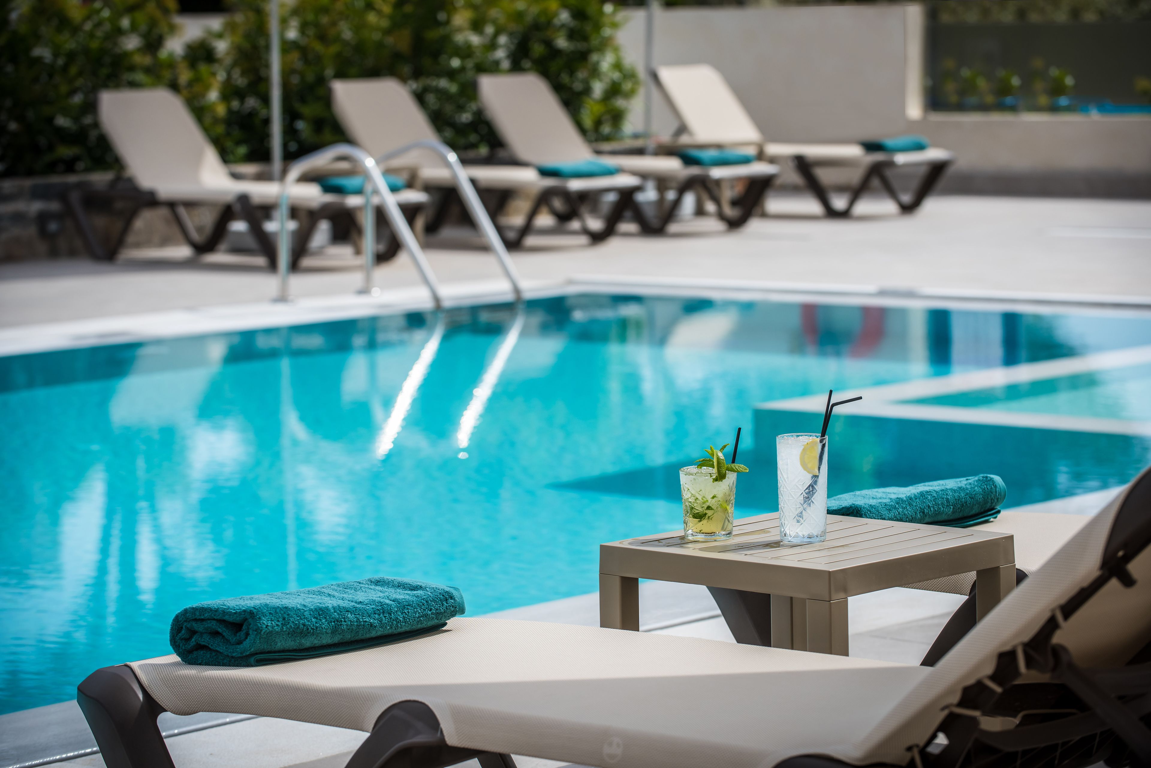 Un área junto a la piscina con sillones de descanso, una mesa con una bebida y una toalla.