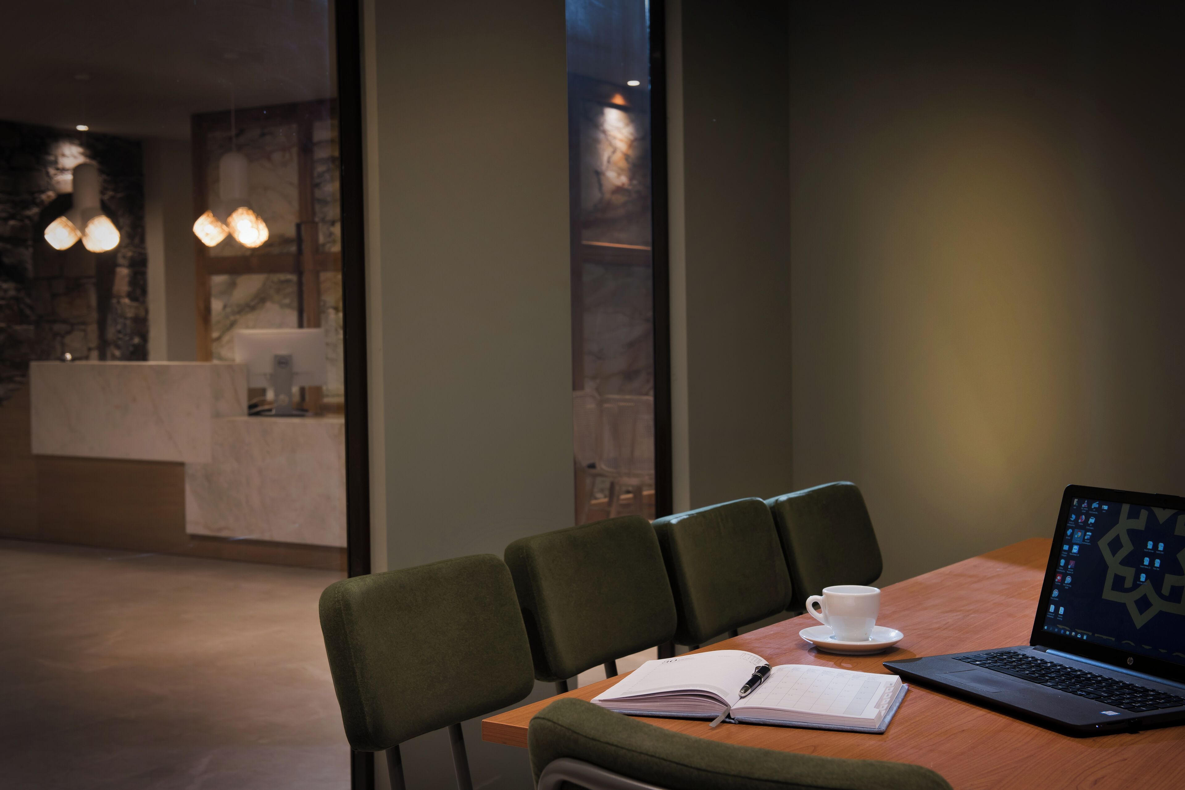 A conference room with a laptop, notebook, and cup on a table, and a reception area in the background.
