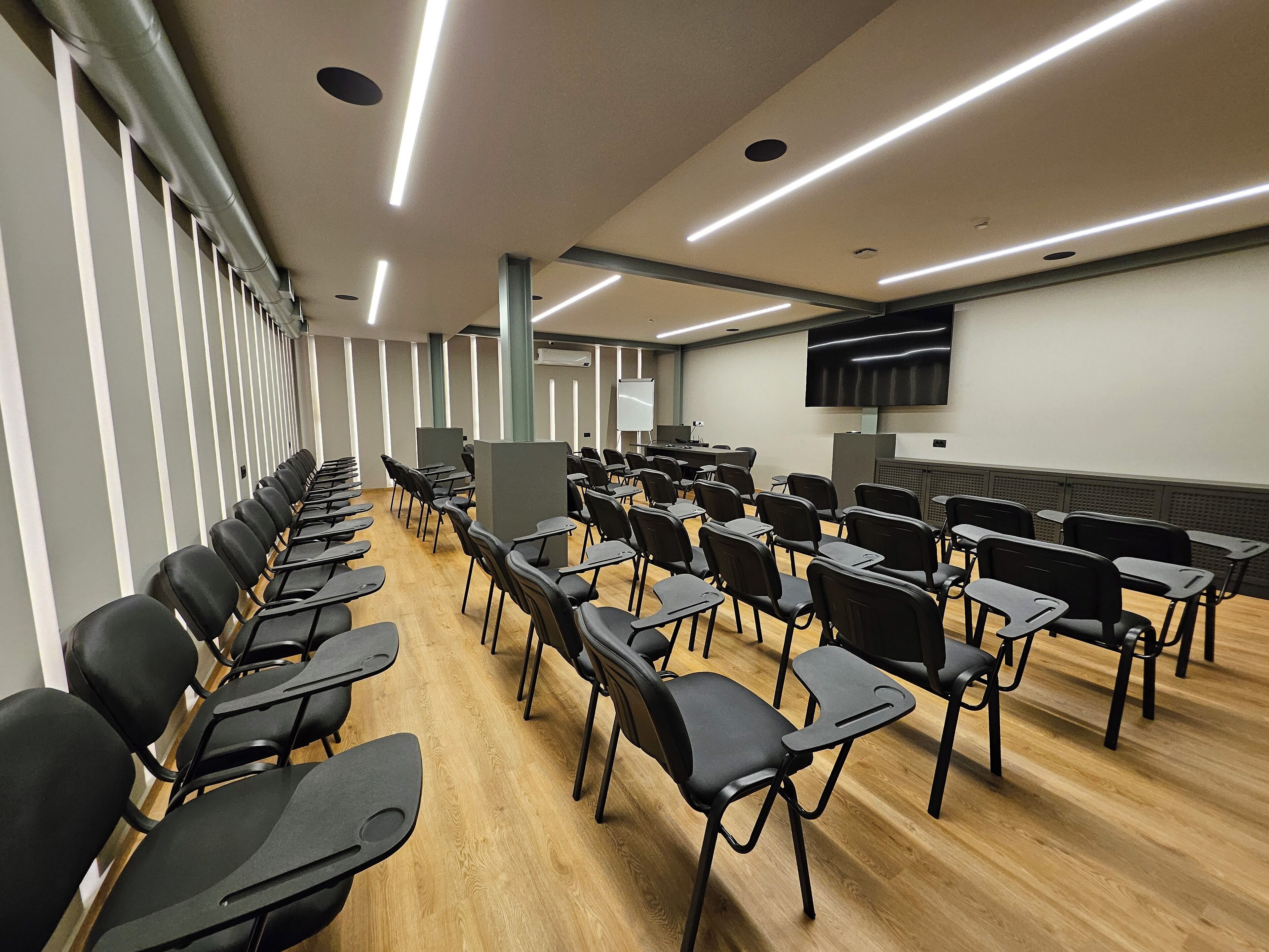 Un salón de conferencias con hileras de sillas negras, piso de madera y una pantalla grande en la pared.