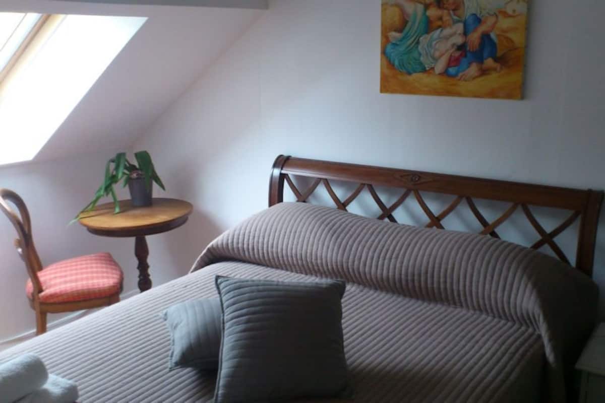 A bedroom with a bed, a chair, a small table with a plant, a lamp, and a painting on the wall.