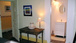 Habitación de hotel con cama, mesita de noche con lámpara, un pequeño cuadro, baño con espejo y lavamanos, y una puerta que da a otra habitación.