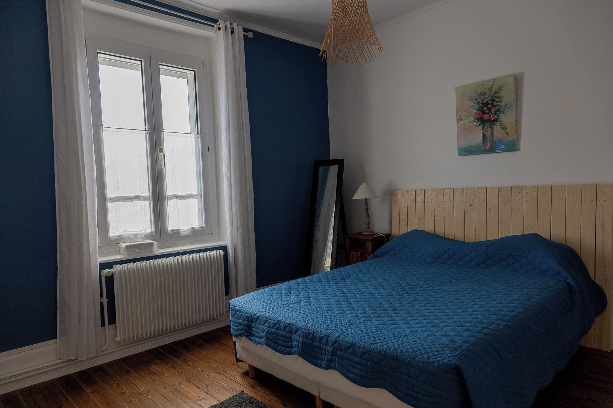 Un dormitorio con cama, ventana con cortinas, radiador, cabecero de madera, mesita de noche con lámpara y un cuadro en la pared.