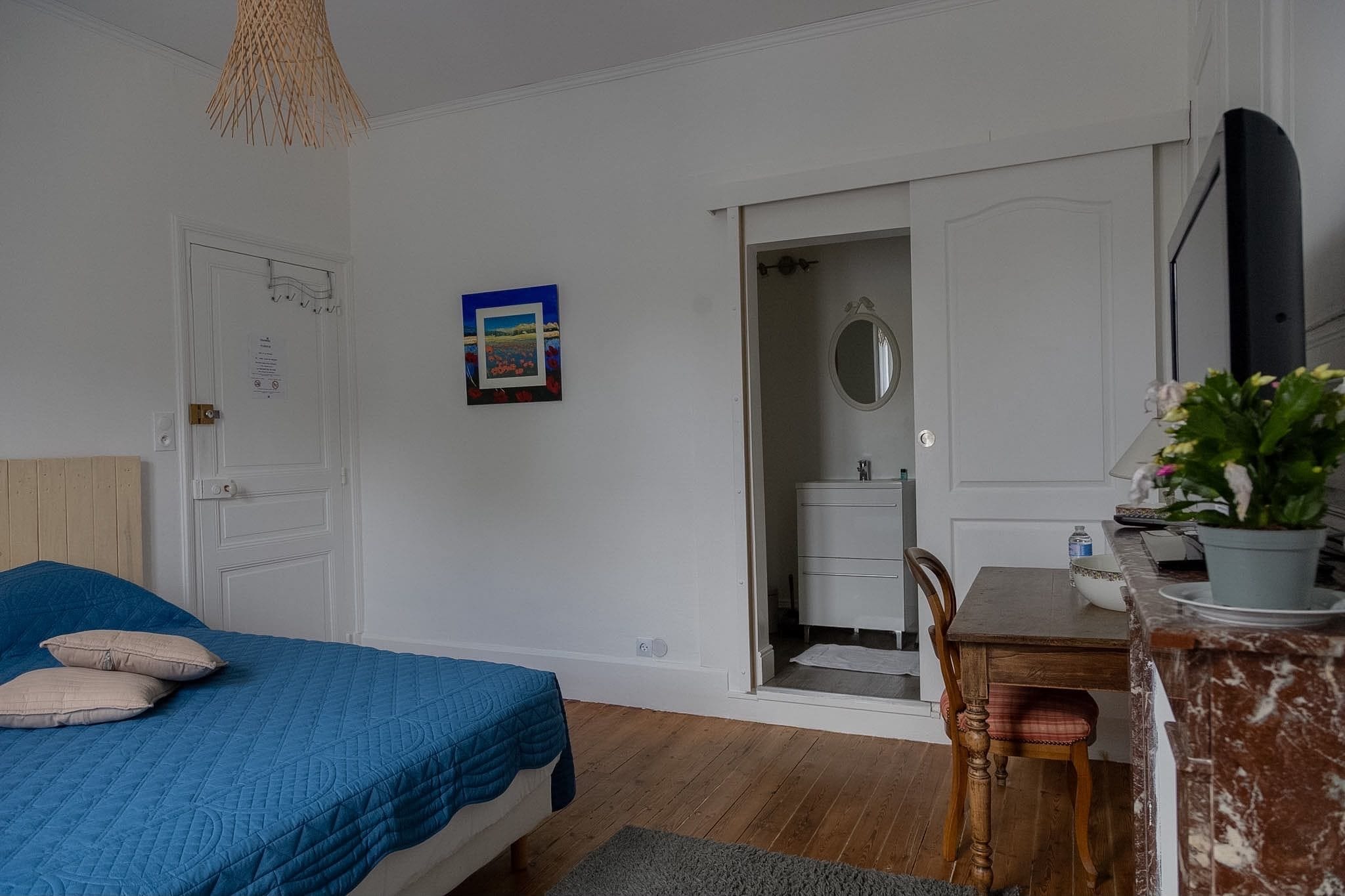 A bedroom with a bed, a desk, a chair, a potted plant, and a door leading to a bathroom.