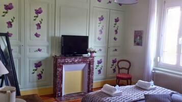 A bedroom with a television on a marble fireplace, a red chair, a bed with towels, and floral wall decorations.