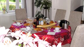 Mesa de comedor preparada con alimentos para el desayuno, como cruasanes y pasteles, en una habitación con sillas y una planta en maceta.