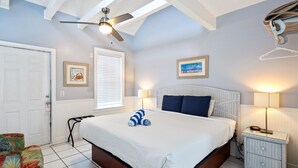 A bedroom with a bed, a ceiling fan, a window with blinds, a framed picture on the wall, and a nightstand with a lamp.