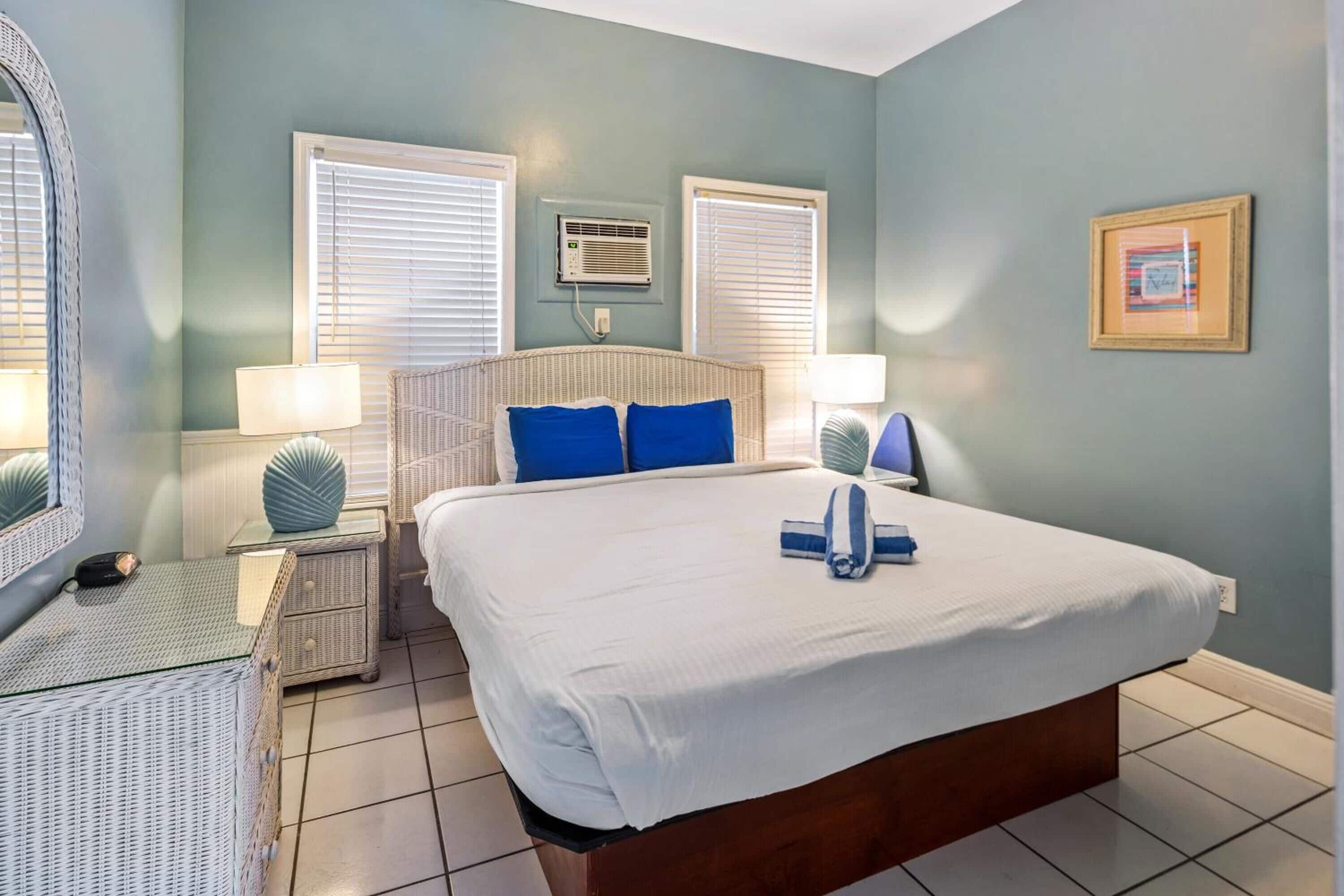 Une chambre à coucher avec un grand lit, deux tables de chevet avec des lampes, un fauteuil en osier et un climatiseur fixé au mur.