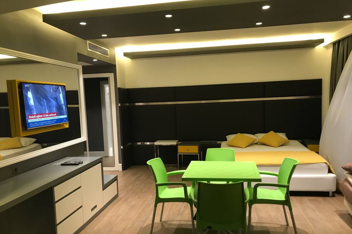 A modern hotel room with a bed, bedside table, TV, green chairs, and a desk.