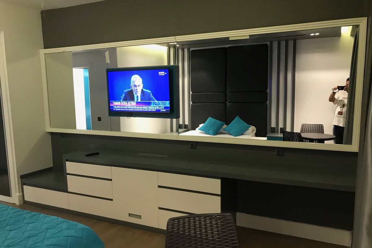 A modern hotel room with a large flat-screen TV mounted on a wall, a black upholstered headboard, a small table with two blue cushions, and a bench.