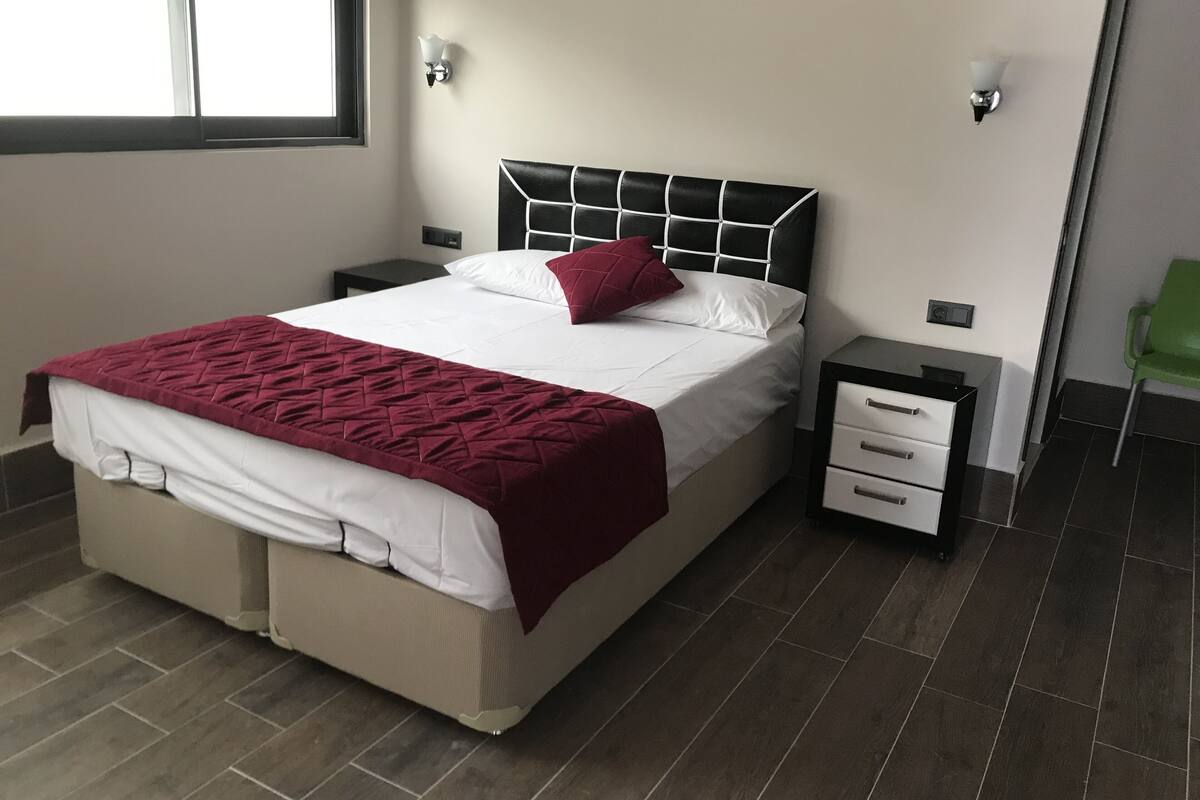 A neatly made bed with a white and maroon bedspread, a black headboard, and two bedside tables with drawers.