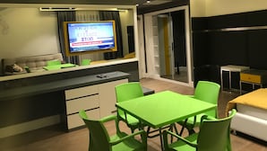 Un espace de vie moderne comprenant un canapé, un téléviseur fixé au mur, une table verte avec des chaises et un lit recouvert d’une couverture jaune.