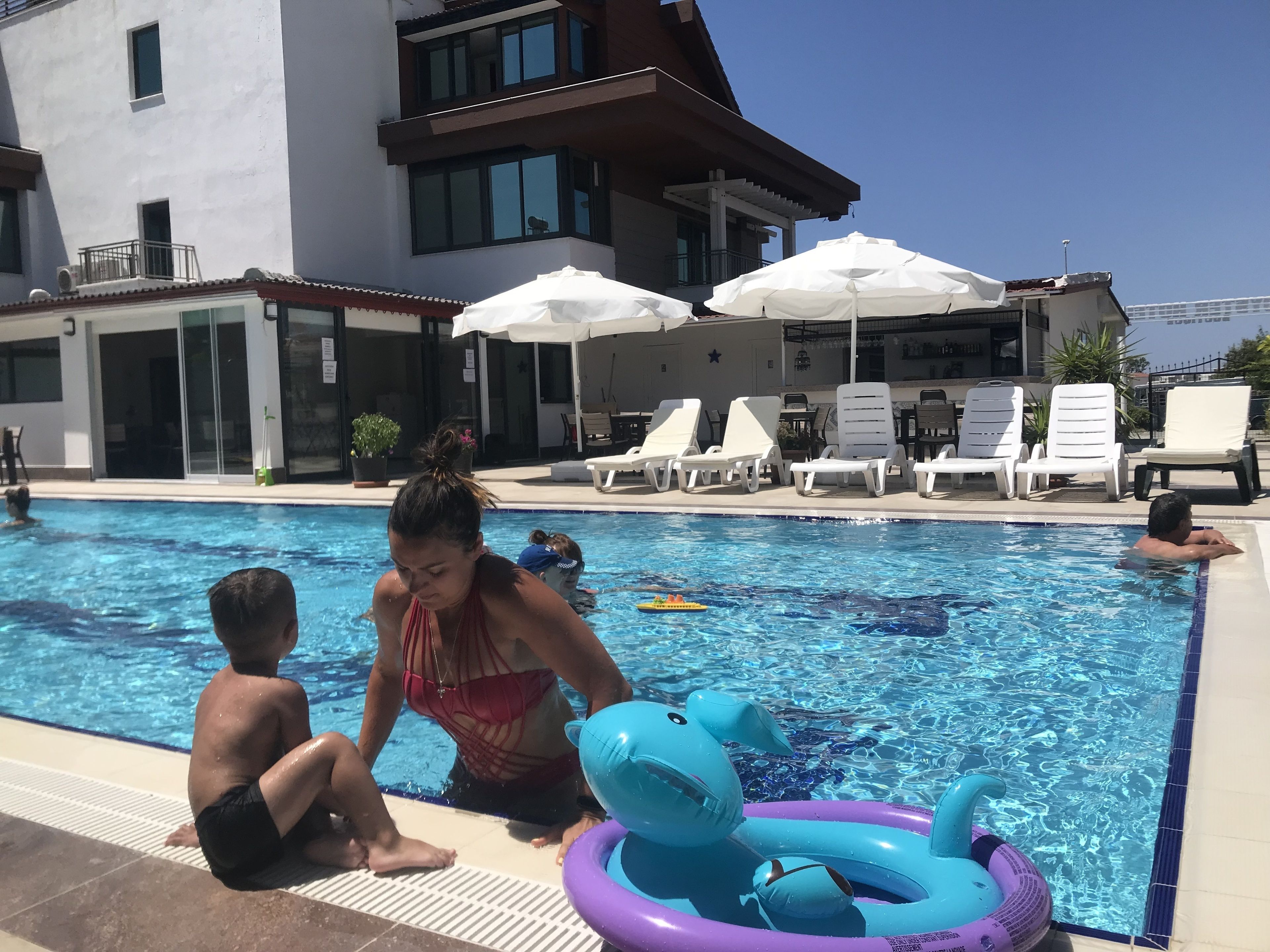 Une piscine avec des chaises longues et des parasols, un bâtiment doté de grandes fenêtres et un ciel d’un bleu limpide.