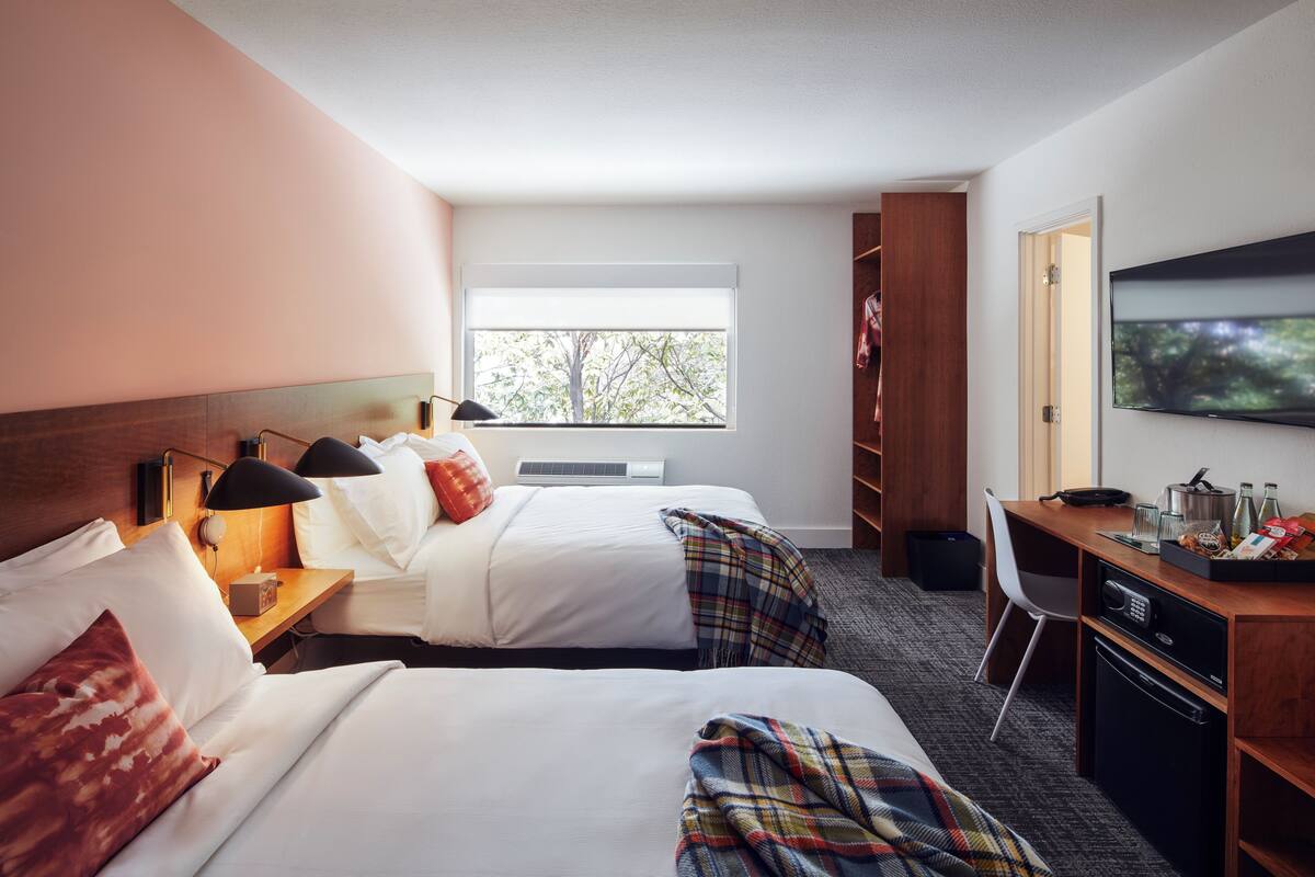 Quarto de hotel com duas camas, uma escrivaninha com micro-ondas, televisão e janela com vista para árvores.