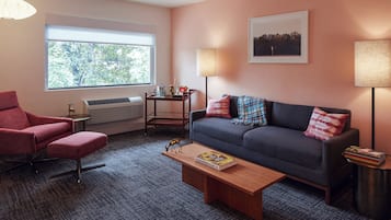 Una sala de estar moderna con un sillón rojo, un sofá gris y una mesa de centro de madera. Se ve un ventanal con vistas a árboles, una lámpara y un cuadro enmarcado en la pared.