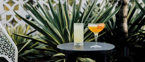 A table with two drinks and a chair in an outdoor setting with a white fence and green plants in the background.