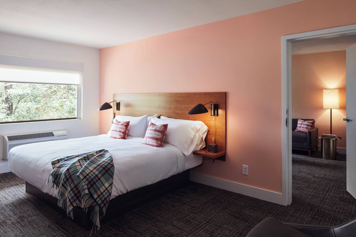 Quarto de hotel com uma cama grande, mesa de cabeceira com abajur, janela com vista para árvores e porta que dá para outro cômodo.