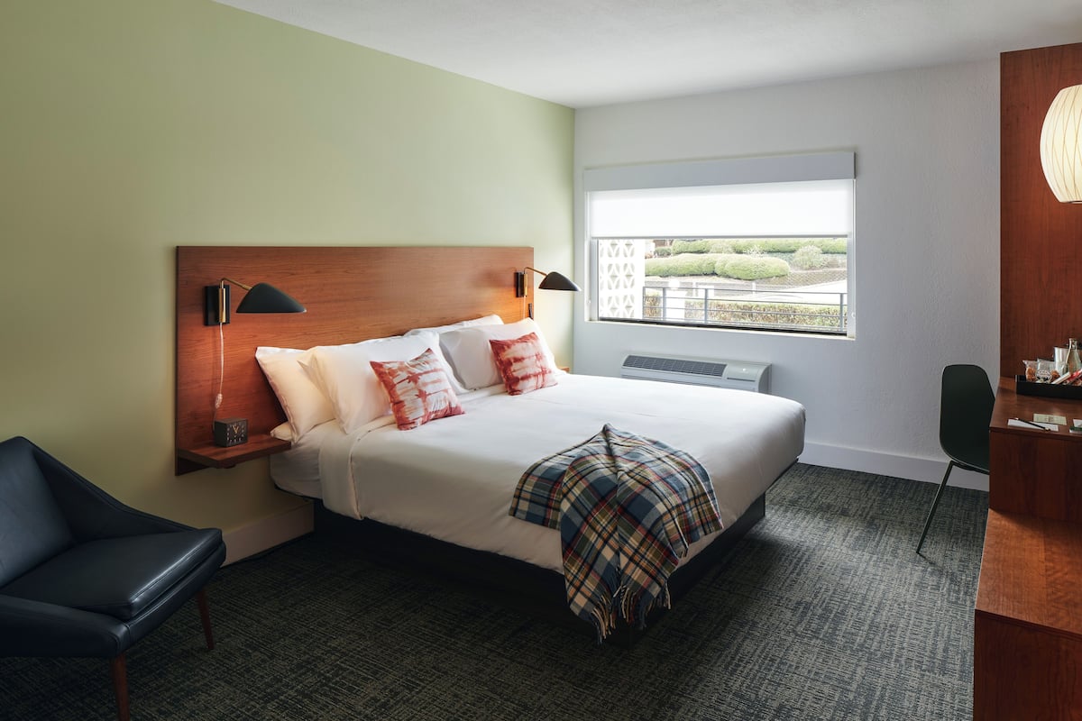 Quarto de hotel com cama, escrivaninha, cadeira, janela com vista e luminária fixada na parede.