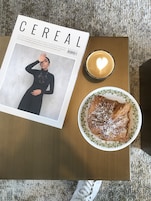 Una revista titulada 'Cereal' sobre una mesa de madera, una taza de café con un diseño de corazón, y un plato de comida.
