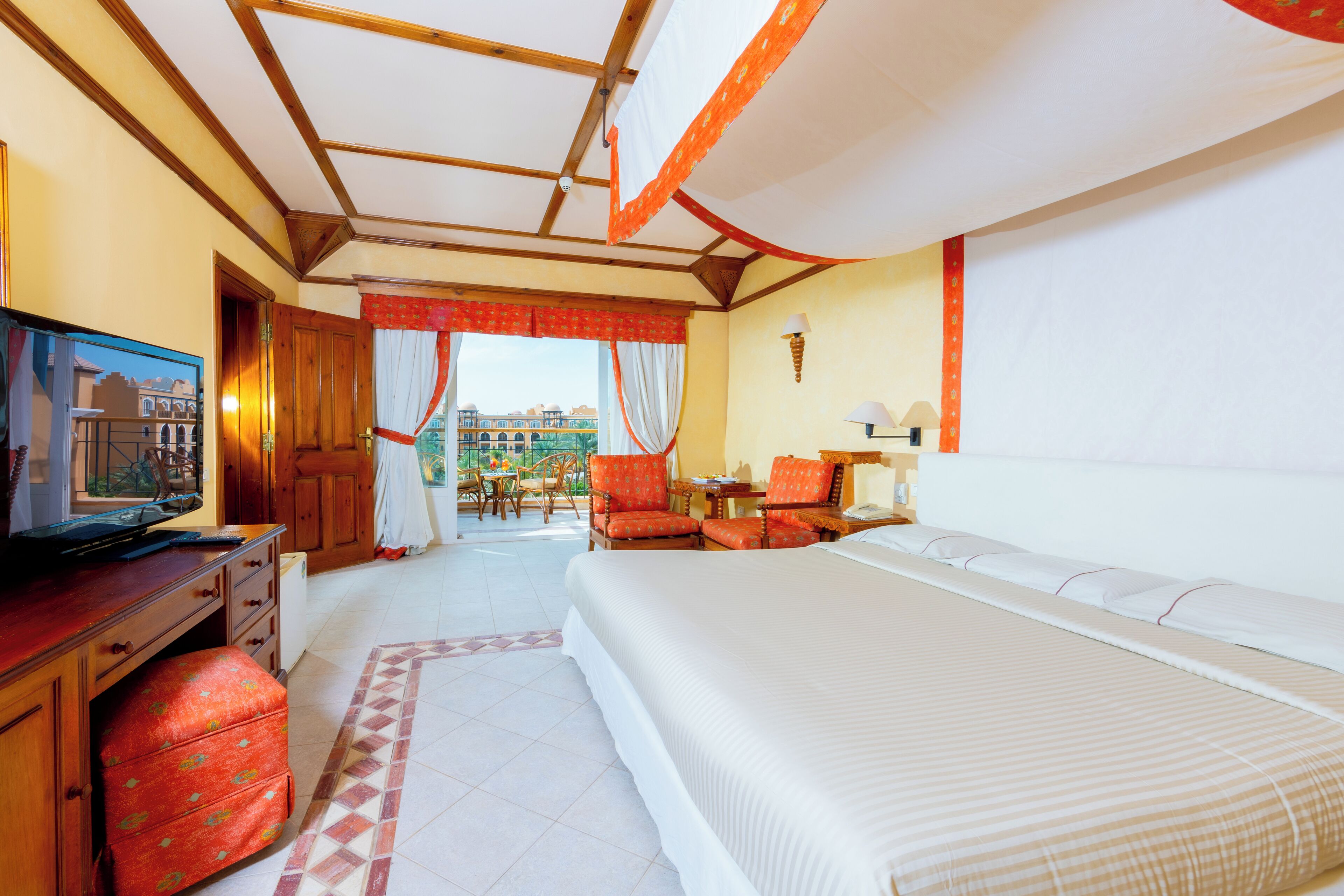 Een hotelkamer met een groot bed, een bureau met een televisie, een klein tafeltje en een stoel. Er is een raam met gordijnen en uitzicht op gebouwen.