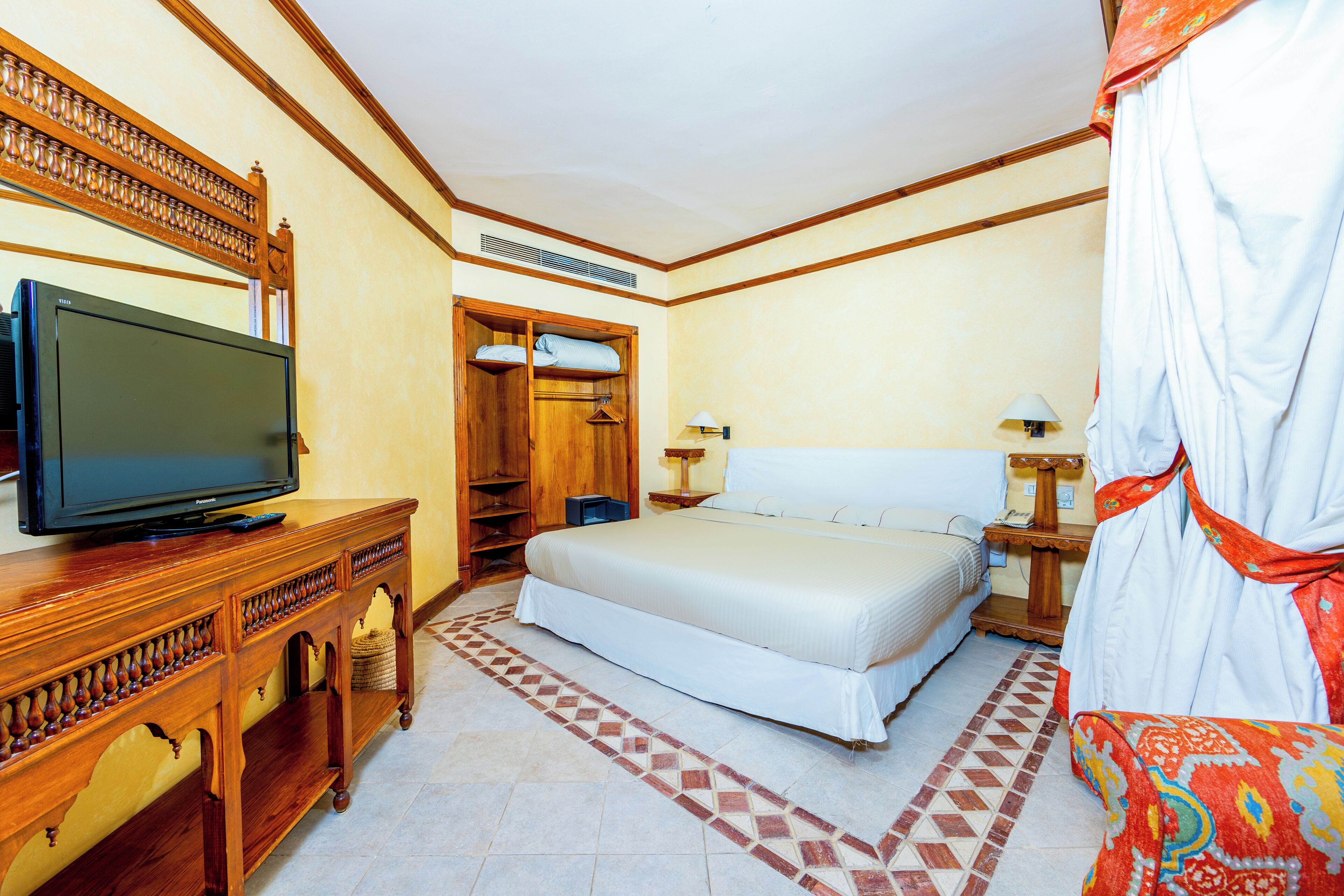 A hotel room with a bed, a television on a wooden cabinet, a wardrobe, a bedside table with a lamp, and a patterned rug.
