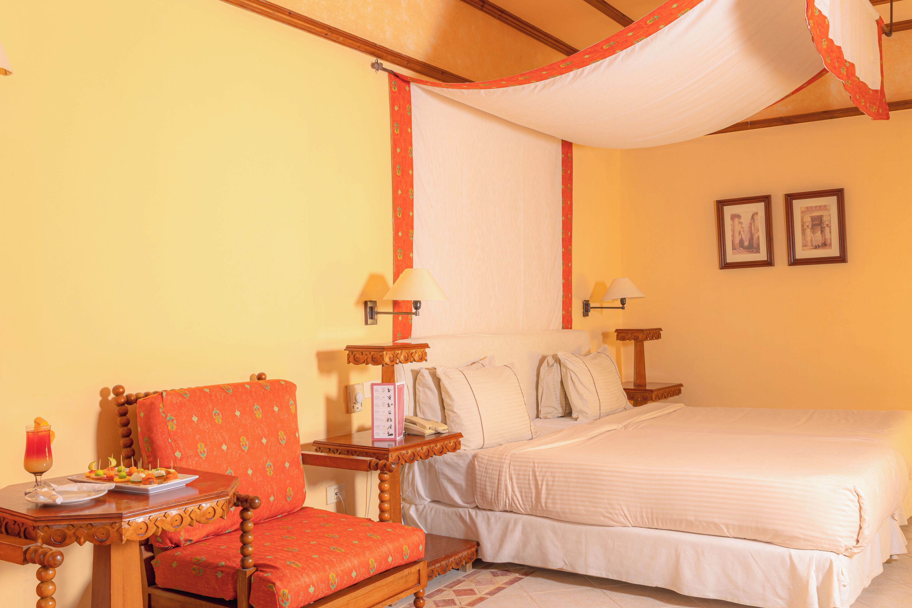 A hotel room with a bed, a chair, a small table with a drink and snacks, and two wall-mounted lamps.