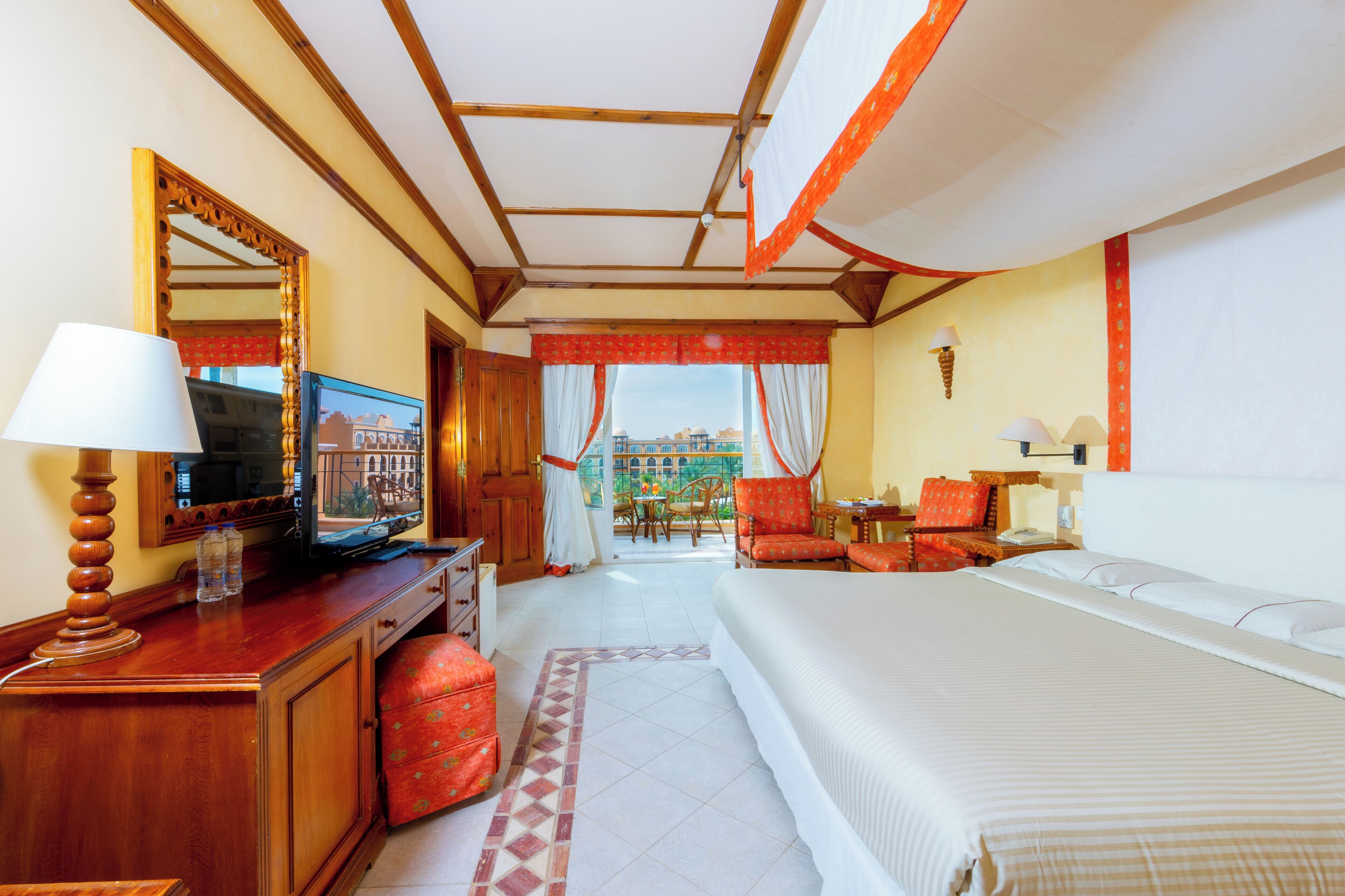 A hotel room with a large bed, a wooden desk with a TV, a mirror, and a view of the outdoors.