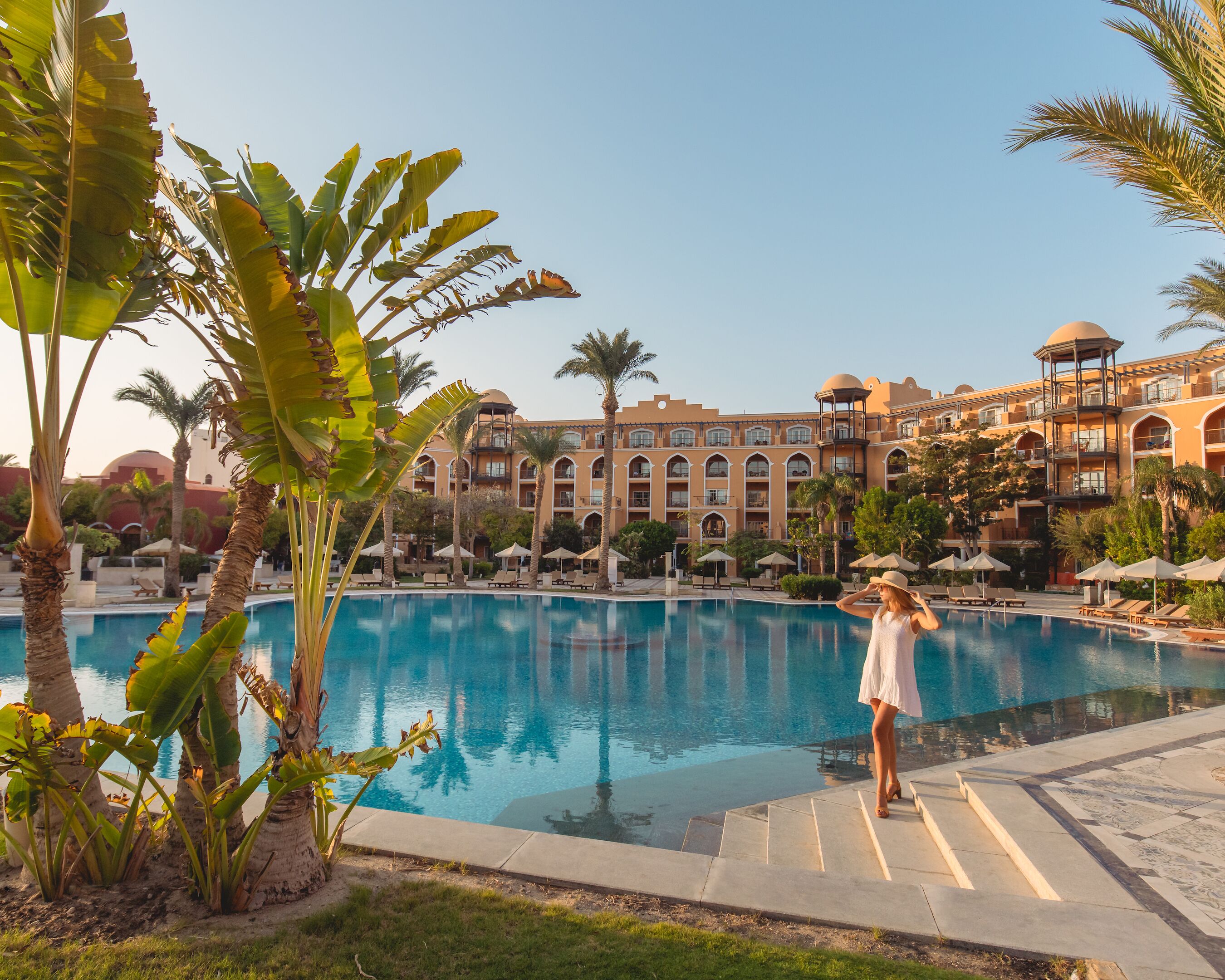 A resort pool with lounge chairs and umbrellas, surrounded by palm trees and a large building with multiple balconies.