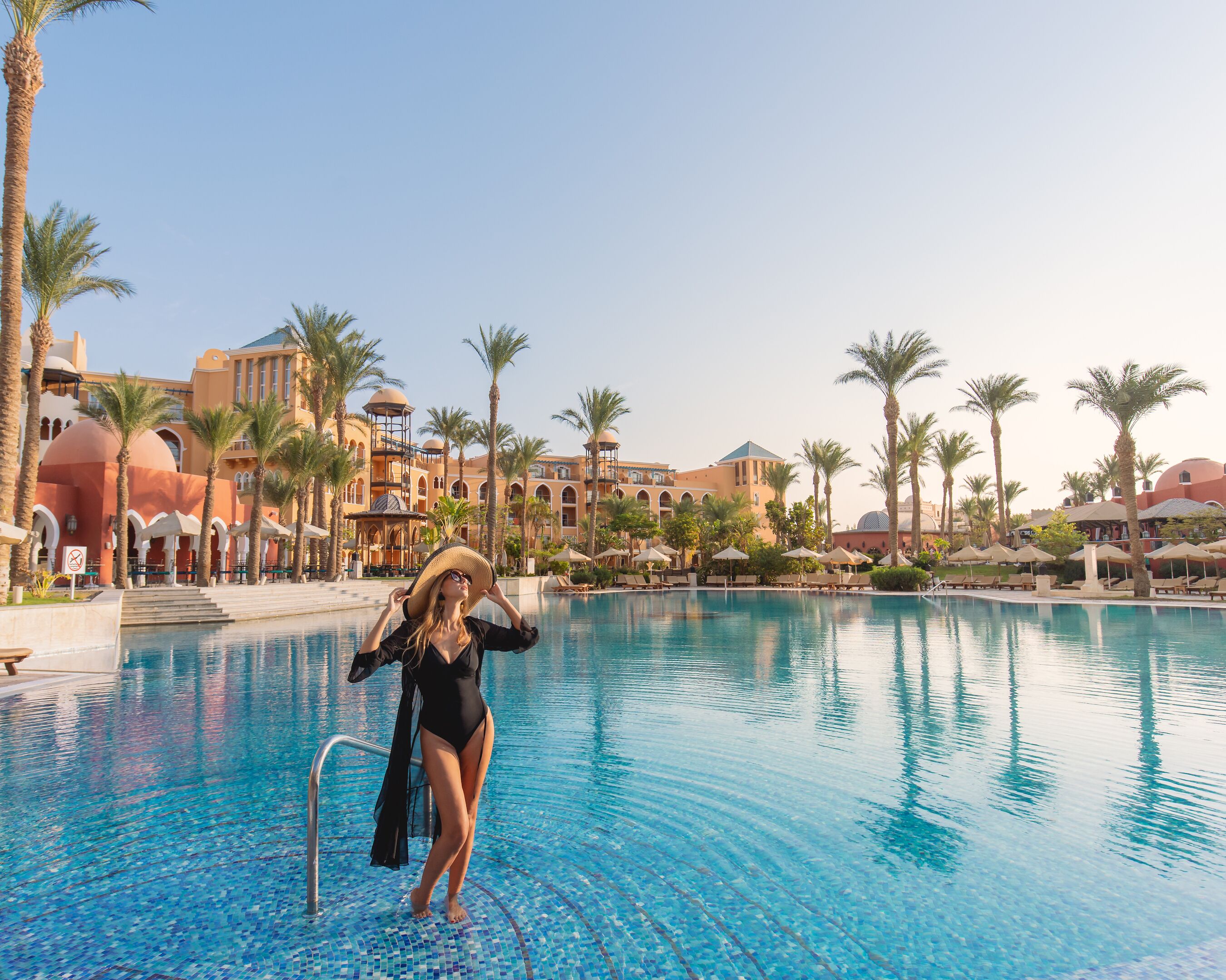 A pool area with a person in a swimsuit, palm trees, and a building with arches and domes in the background.