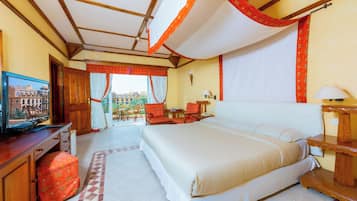 A hotel room with a large bed, a television, a desk with a computer, and a balcony with a view of buildings.