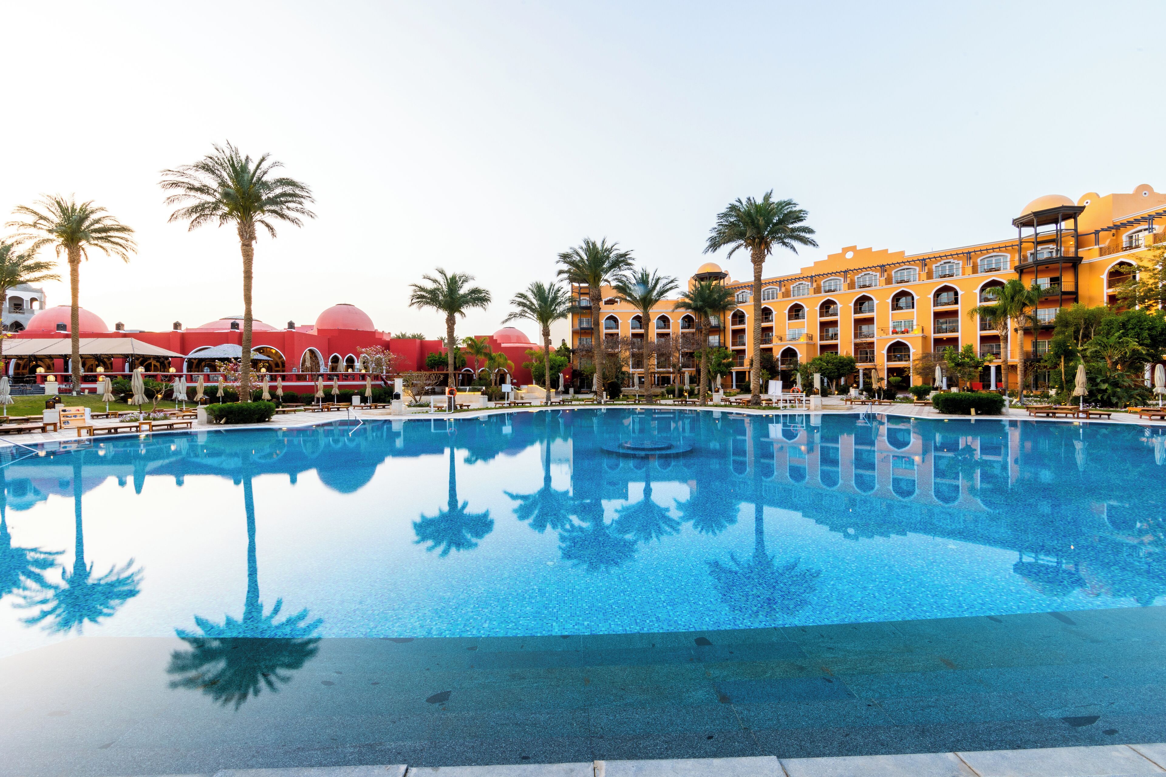 Una piscina grande rodeada de palmeras y un hotel con varios balcones y ventanas arqueadas.