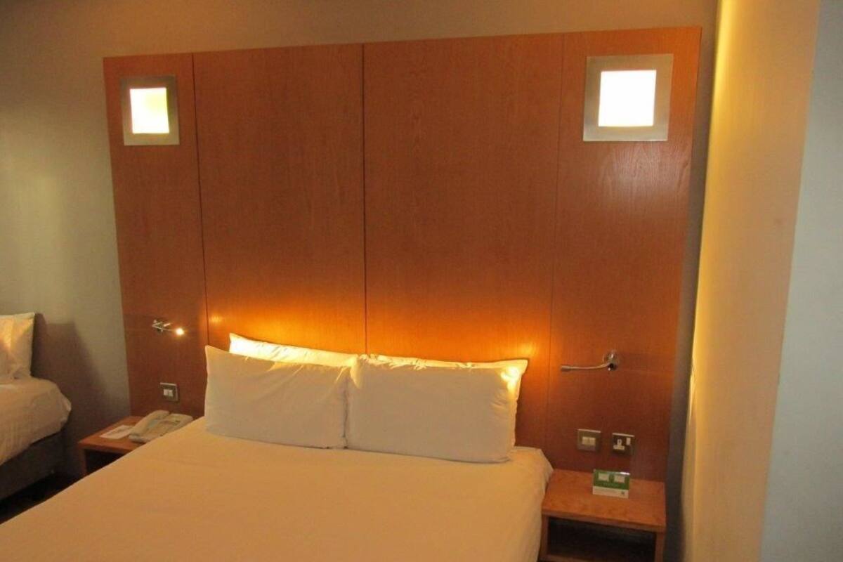 A hotel room with a large bed, two bedside tables, a nightstand, and a wooden headboard with built-in lighting.