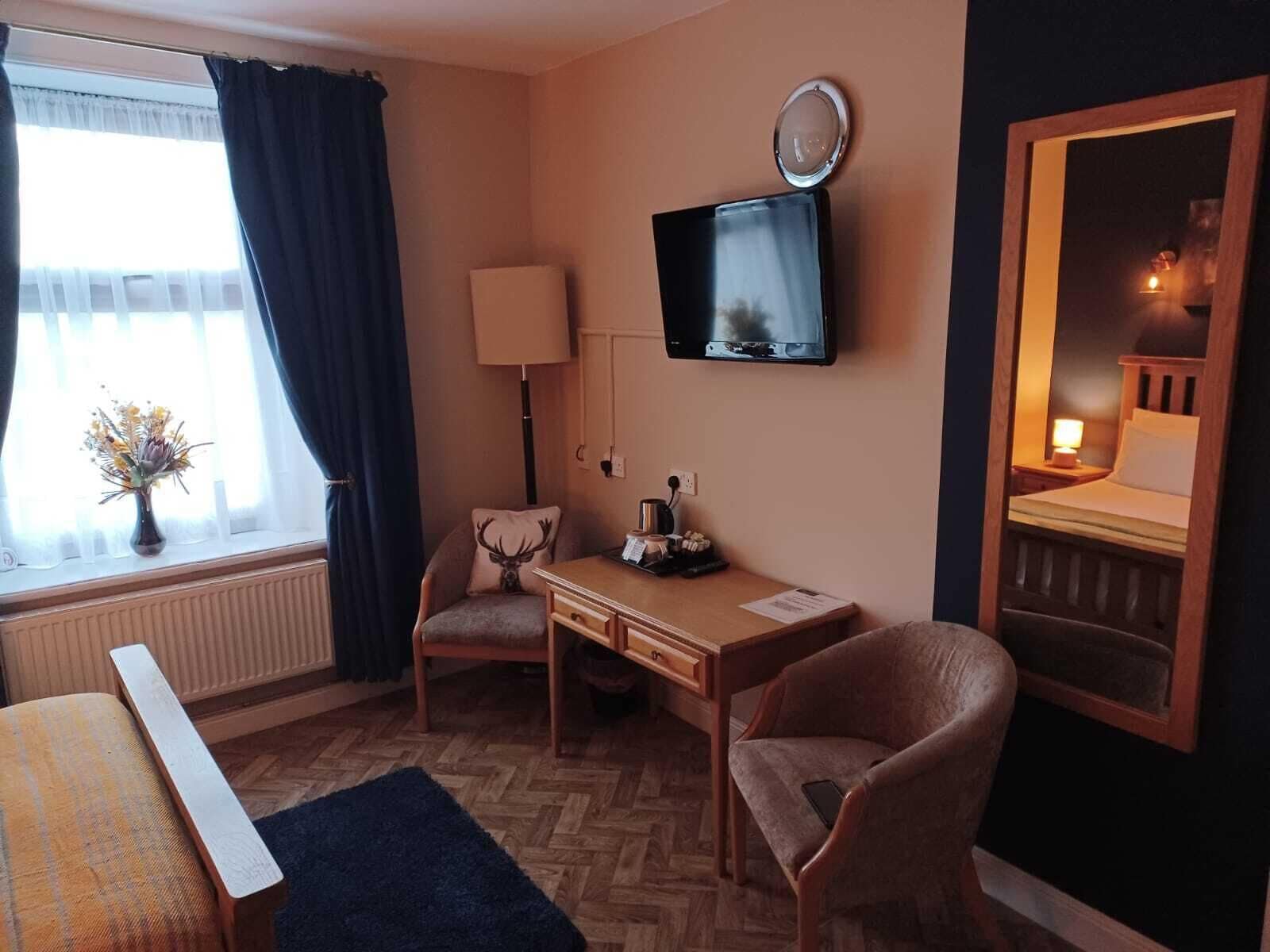 A hotel room with a wooden desk, two chairs, a television, a mirror, a window with curtains, and a radiator.