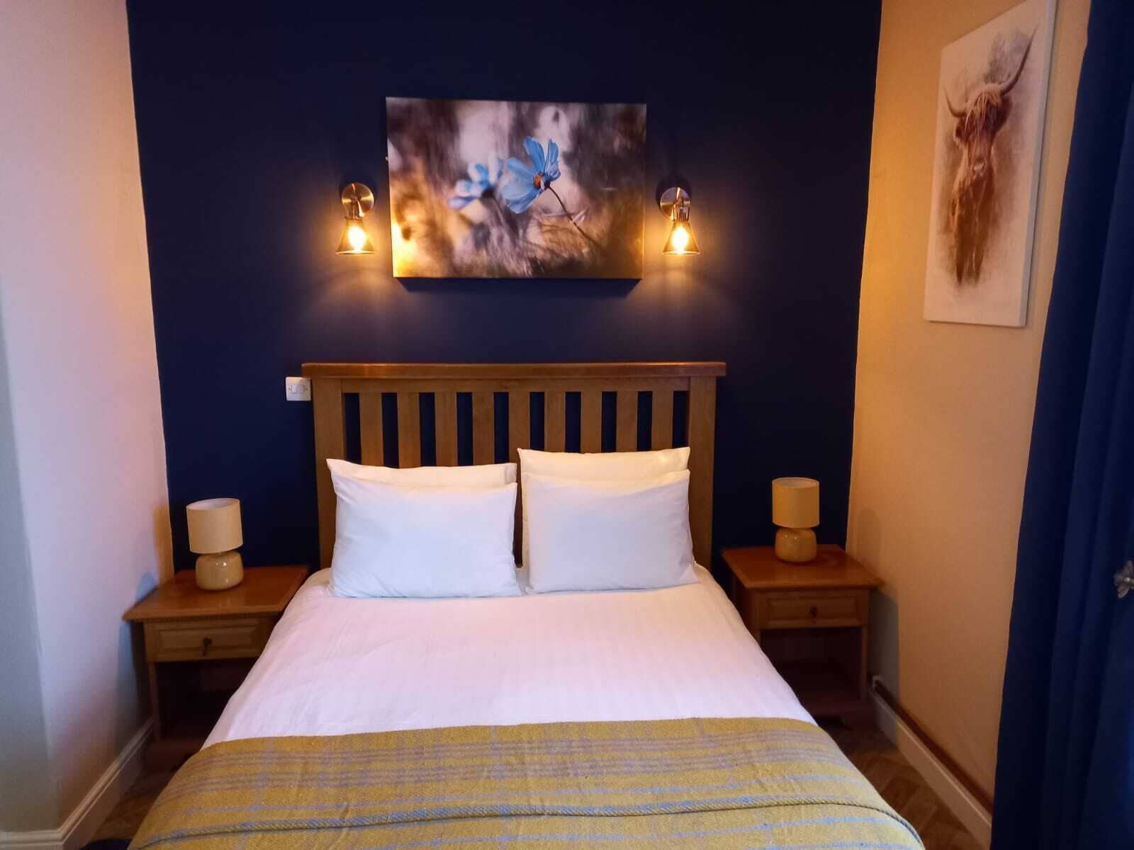 Ein Hotelzimmer mit einem hölzernen Kopfteil, zwei weißen Kissen, einem Bett mit einer gelb-karierten Decke, einem Gemälde über dem Bett und zwei Nachttischlampen.