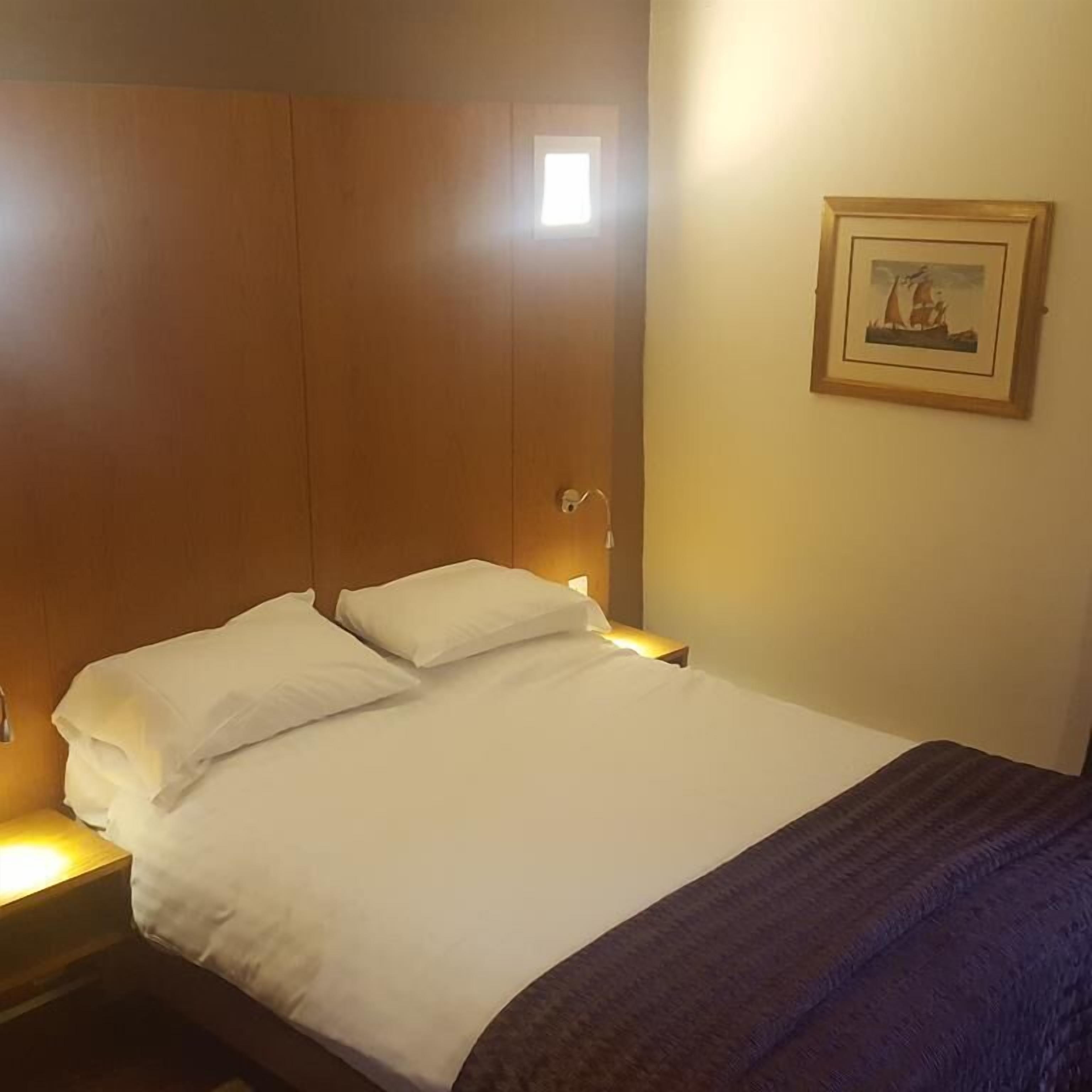 Een hotelkamer met een bed, nachtkastje, wandlamp en een lijst met een afbeelding.