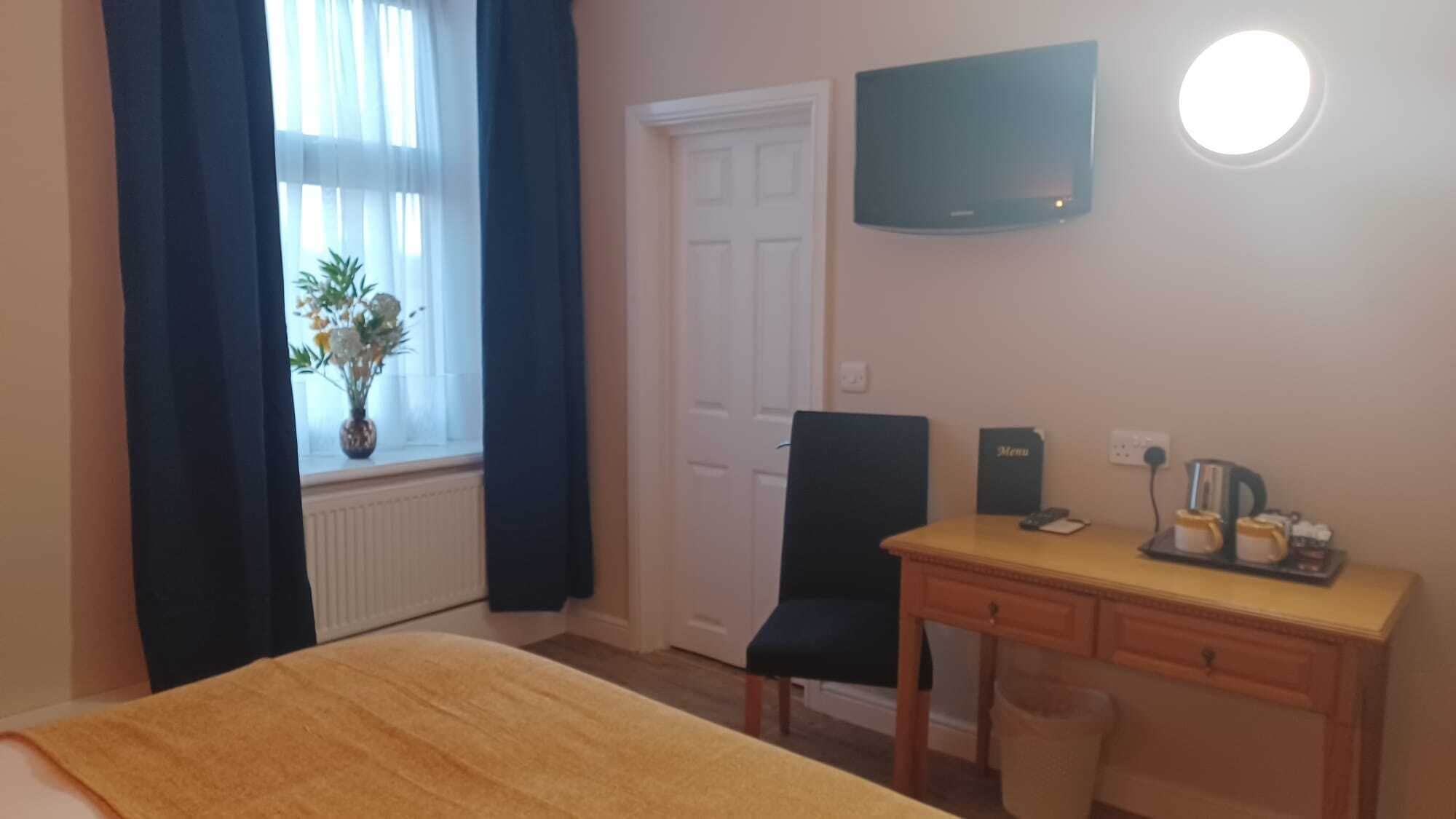 Quarto de hotel com cama, escrivaninha com cadeira, televisão na parede e janela com cortinas.