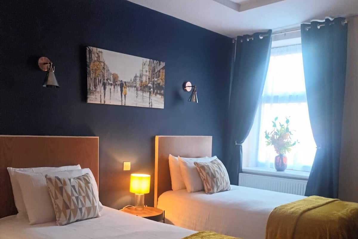 A hotel room with two beds, a nightstand with a lamp, a wall-mounted clock, a painting, and a window with curtains.