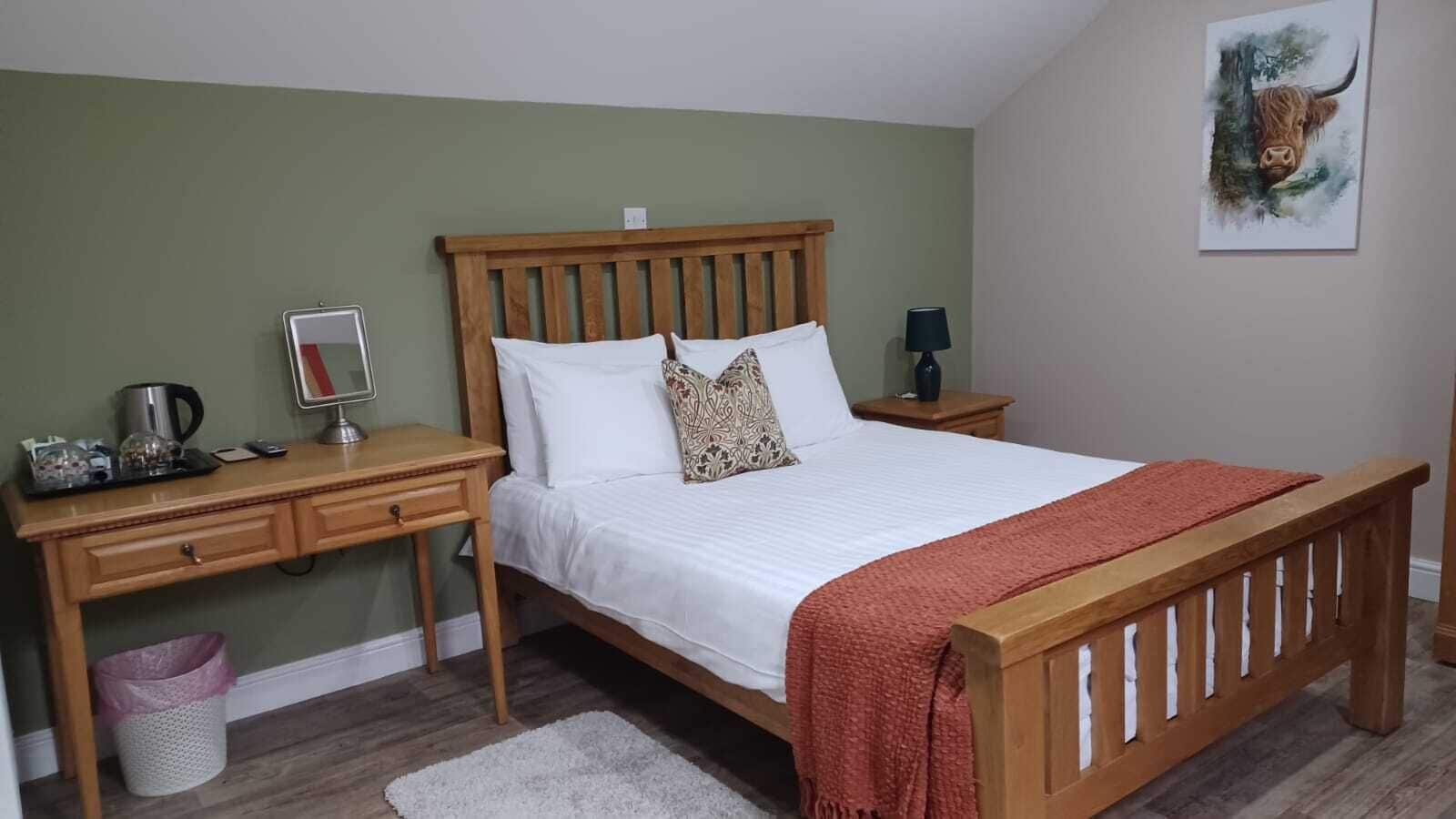 A bedroom with a wooden bed, a desk, a chair, a bedside table, a lamp, and a painting on the wall.