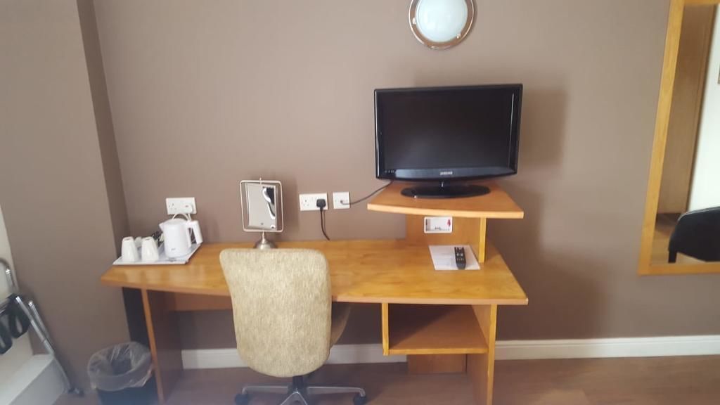 Een kamer met een houten bureau, een zwarte flatscreen-tv, een stoel, een prullenbak en een spiegel bevestigd aan de muur.