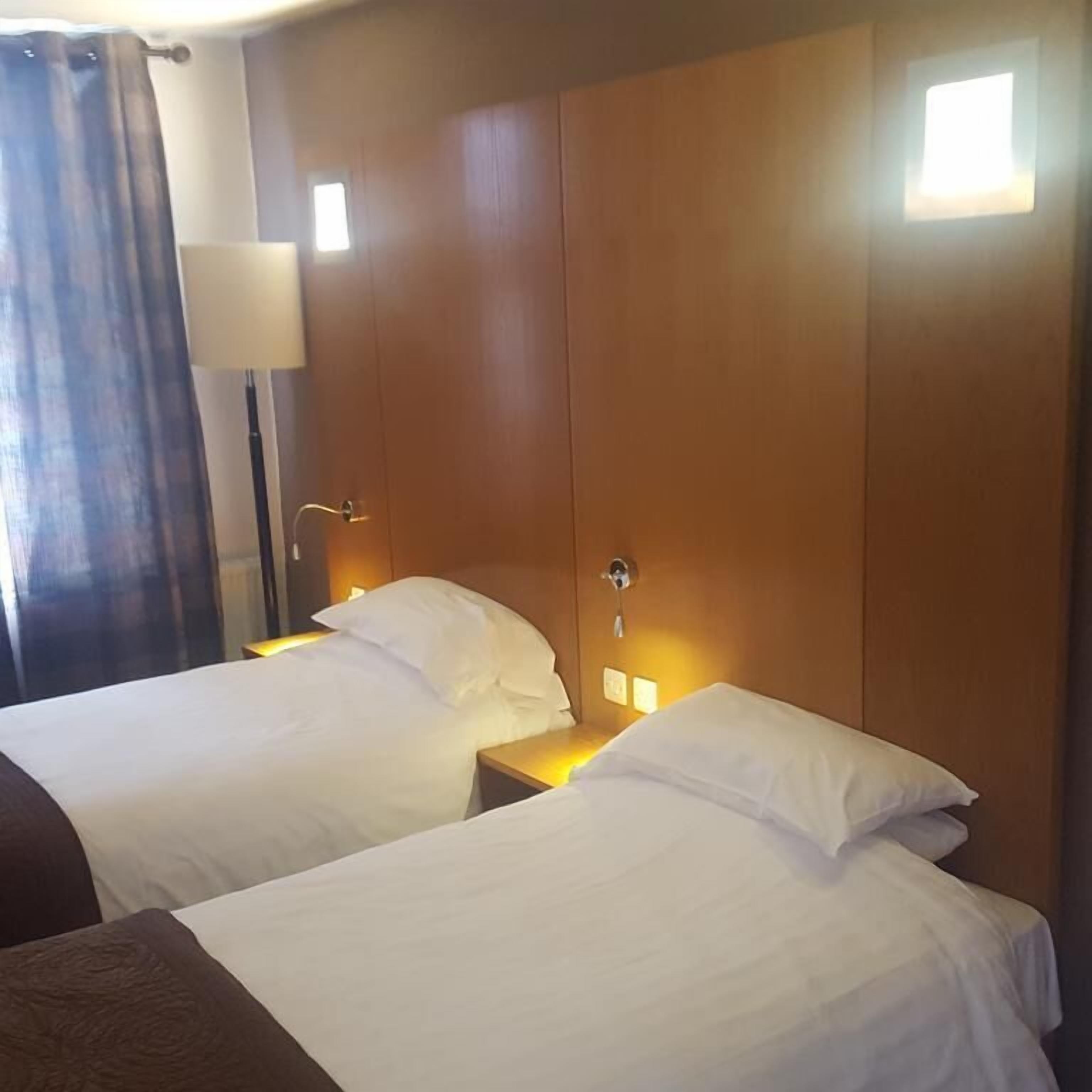 A hotel room with two beds, a nightstand with a lamp, and a window with curtains.
