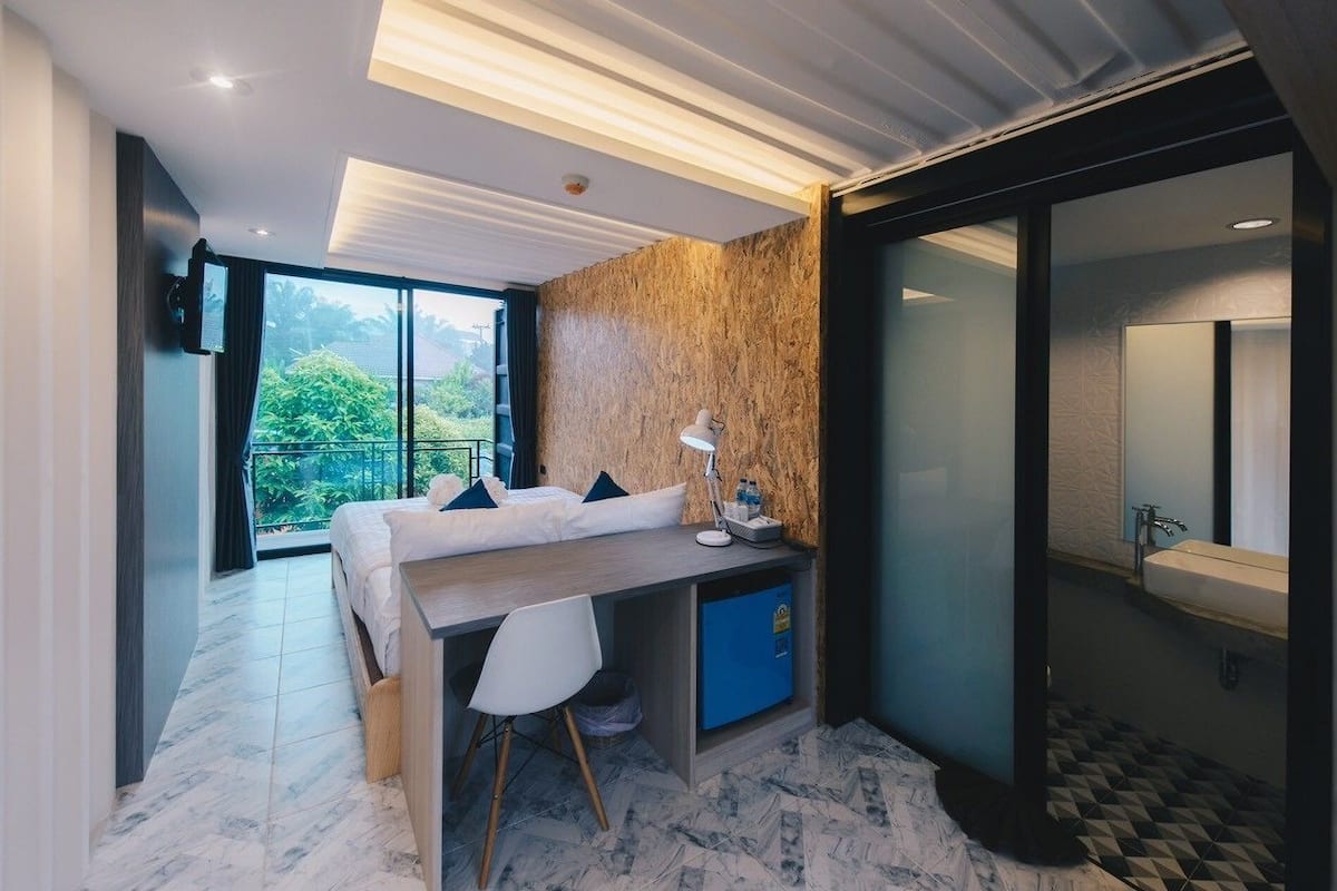 Un dormitorio moderno con cama, escritorio, silla y un amplio ventanal que ofrece vistas a zonas verdes.