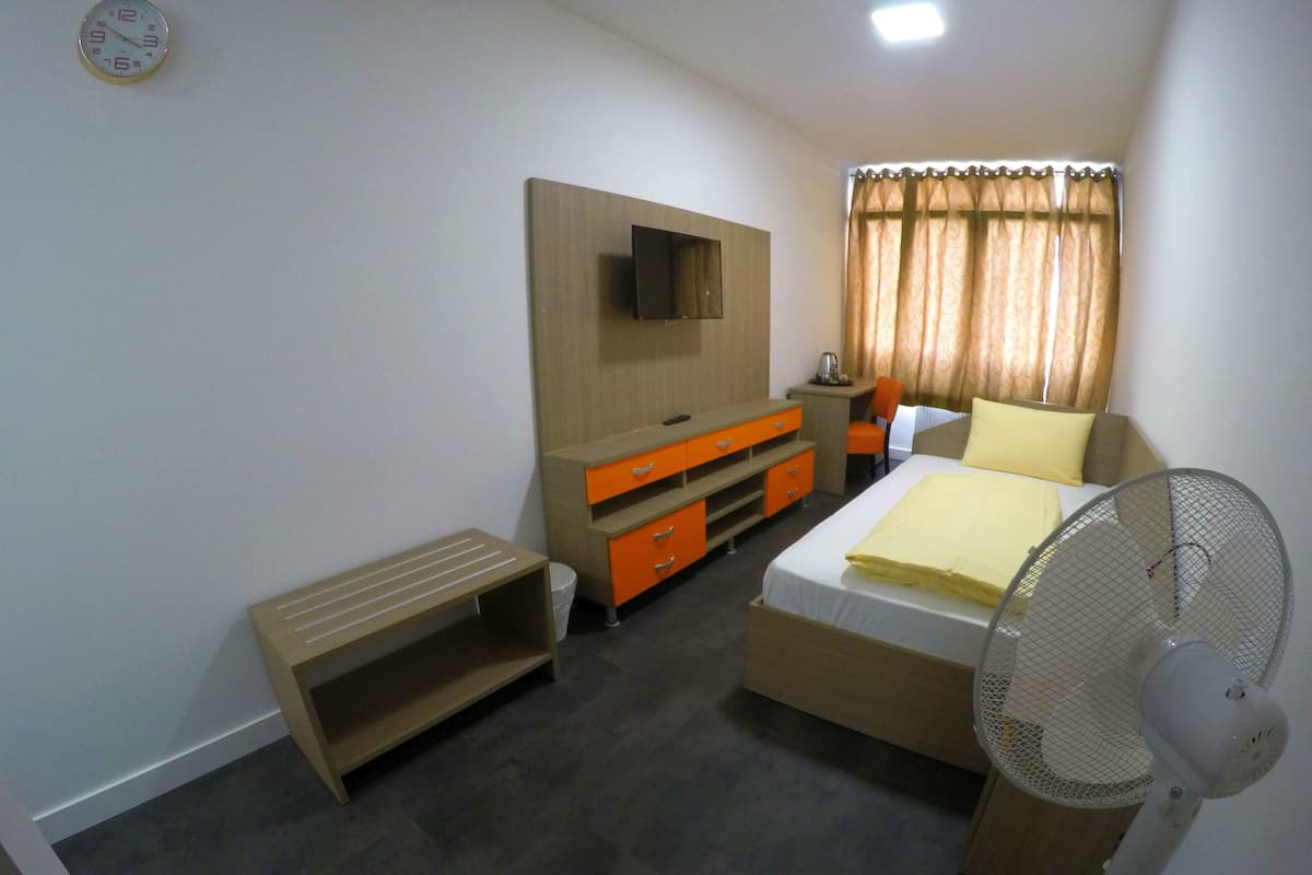 A bedroom with a bed, a desk, a chair, a TV mounted on the wall, and a standing fan.