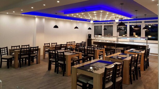 A modern restaurant interior with multiple dining tables and chairs, a central counter with a display case, and blue ambient lighting on the ceiling. - Terminal Hotel One (Sindelfingen)
