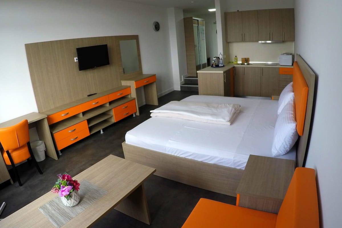 A hotel room with a bed, a desk with a chair, a TV, a small table with a vase of flowers, and a kitchenette.